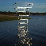 Une sculpture géométrique en métal de Clara Imbert avec quatre cadres rectangulaires empilés se dresse dans une eau peu profonde près d'un rivage herbeux, se reflétant à la surface - un exemple frappant de ses techniques sculpturales sous un ciel clair.
