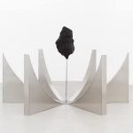 Un grand rocher sombre de forme irrégulière, évoquant les techniques de Clara Imbert, est suspendu au-dessus du sol sur un poteau métallique, entouré de quatre panneaux métalliques argentés incurvés disposés symétriquement sur un sol et un fond blancs.