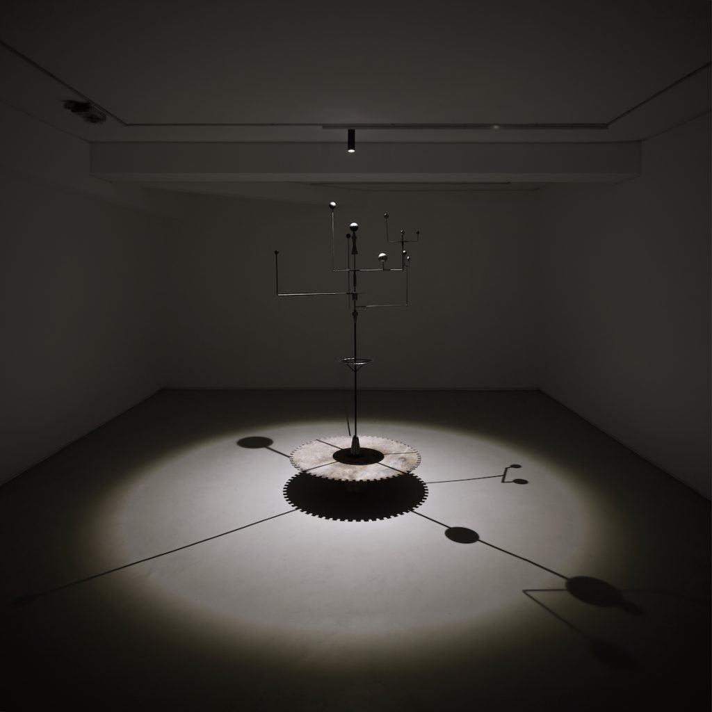 Une sculpture cinétique en métal de Clara Imbert trône au centre d'une pièce faiblement éclairée, projetant de multiples ombres sur le sol. Composée d'une tige centrale verticale, elle est pourvue de divers bras horizontaux et de formes géométriques, mettant en valeur des techniques innovantes.