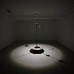 Une sculpture cinétique en métal de Clara Imbert trône au centre d'une pièce faiblement éclairée, projetant de multiples ombres sur le sol. Composée d'une tige centrale verticale, elle est pourvue de divers bras horizontaux et de formes géométriques, mettant en valeur des techniques innovantes.