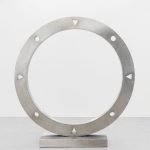 Une grande sculpture circulaire en métal de Clara Imbert repose sur une base rectangulaire. L'anneau présente des trous circulaires régulièrement espacés et des découpes triangulaires sur son pourtour, mettant en valeur des techniques uniques sur un fond uni et clair.