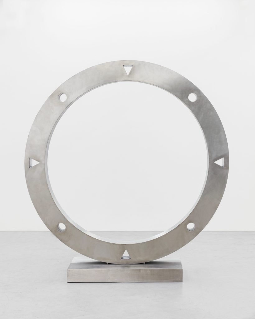 Une grande sculpture circulaire en métal de Clara Imbert repose sur une base rectangulaire. L'anneau présente des trous circulaires régulièrement espacés et des découpes triangulaires sur son pourtour, mettant en valeur des techniques uniques sur un fond uni et clair.