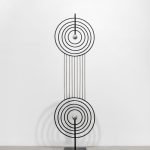 Une sculpture en métal de Clara Imbert, composée de deux motifs circulaires concentriques reliés verticalement par des tiges droites, chaque cercle contenant une sphère métallique centrale, met en valeur des techniques et des expressions saisissantes. Montée sur un socle rectangulaire noir sur fond blanc.