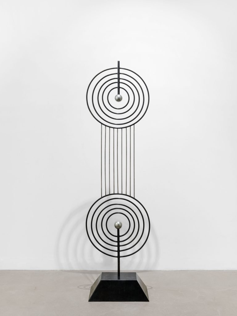Une sculpture en métal de Clara Imbert, composée de deux motifs circulaires concentriques reliés verticalement par des tiges droites, chaque cercle contenant une sphère métallique centrale, met en valeur des techniques et des expressions saisissantes. Montée sur un socle rectangulaire noir sur fond blanc.