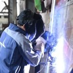 Une personne portant un casque de soudage et des gants soude du métal dans un atelier industriel. Des étincelles et une lumière vives, issues du procédé de brouillon automatique, sont visibles au point de soudure.