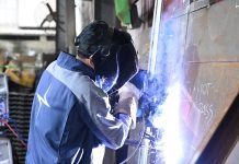 Une personne portant un casque de soudage et des gants soude du métal dans un atelier industriel. Des étincelles et une lumière vives, issues du procédé de brouillon automatique, sont visibles au point de soudure.