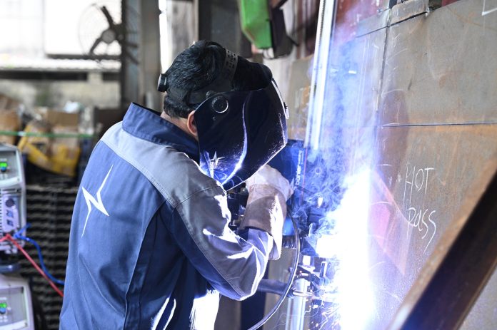 Une personne portant un casque de soudage et des gants soude du métal dans un atelier industriel. Des étincelles et une lumière vives, issues du procédé de brouillon automatique, sont visibles au point de soudure.