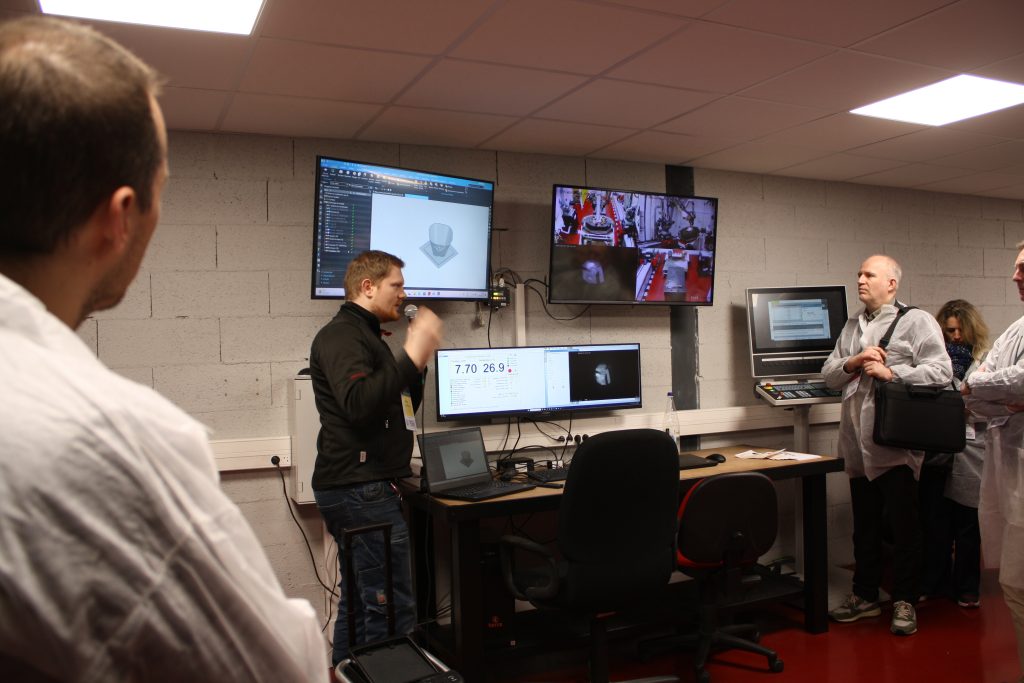 Un groupe de personnes se tient dans une salle aux murs de briques grises et observe une présentation lors de la journée technique. Plusieurs écrans diffusent des graphiques et des vidéos numériques. Une personne à un bureau explique par des gestes, tandis que d'autres écoutent attentivement, certains portant des blouses de laboratoire Irepa Laser.