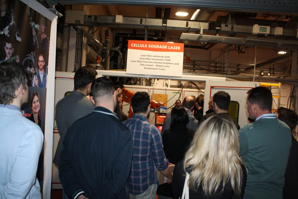 Un groupe de personnes se tient devant un poste de soudage laser surmonté d'une enseigne lors d'une journée technique chez Irepa Laser. La salle industrielle, remplie de machines, accueille une foule attentive, observant ou participant peut-être à une démonstration de fusion.