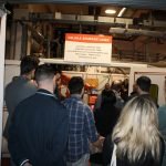 Un groupe de personnes se tient devant un poste de soudage laser surmonté d'une enseigne lors d'une journée technique chez Irepa Laser. La salle industrielle, remplie de machines, accueille une foule attentive, observant ou participant peut-être à une démonstration de fusion.