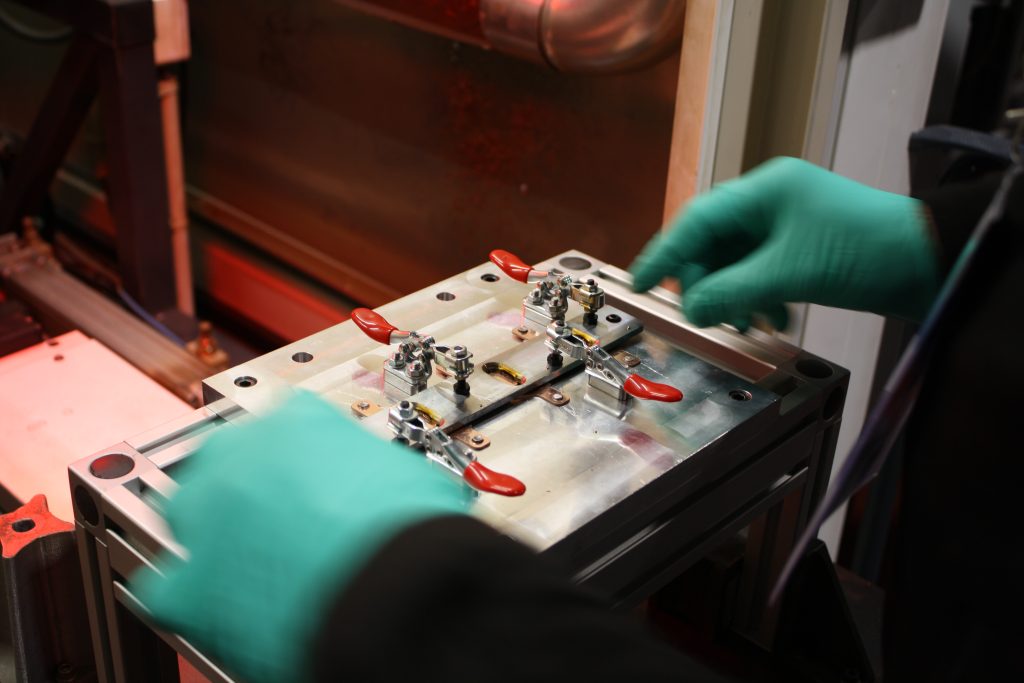 Lors de la première édition réussie de notre journée technique, une personne portant des gants verts manipule avec précaution une pièce métallique complexe aux poignées rouges. L'atelier Laser Fusion d'Irepa bourdonne en arrière-plan, flou mais animé par ses équipements et ses lumières.