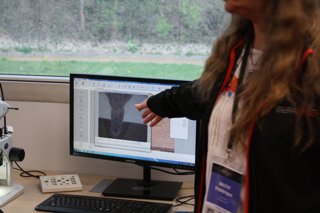 Une personne fait un geste vers un écran d'ordinateur affichant un gros plan d'un matériau, rappelant la technique de la Journée. Un microscope est posé sur le bureau, à côté de l'écran. La fenêtre derrière révèle la verdure à l'extérieur, rappelant la première édition couronnée de succès.