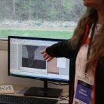 Une personne fait un geste vers un écran d'ordinateur affichant un gros plan d'un matériau, rappelant la technique de la Journée. Un microscope est posé sur le bureau, à côté de l'écran. La fenêtre derrière révèle la verdure à l'extérieur, rappelant la première édition couronnée de succès.