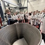 Un groupe de personnes en blouse blanche écoute attentivement un conférencier lors d'une journée technique. Au premier plan, un grand objet imprimé en 3D, partiellement construit, trône. Le décor industriel met en scène des machines et équipements d'Irepa Laser en arrière-plan.