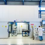 Un technicien en blouse blanche utilise un panneau de commande devant une grande machine laser à procédé, dans une installation moderne et lumineuse. Des écrans et des panneaux d'avertissement sont visibles sur et autour de l'équipement.