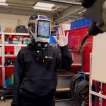 Une personne portant un casque de soudeur et un sweat à capuche noir se tient à l'intérieur, une main levée, montrant un motif bleu Brouillon sur la paume. Des équipements industriels, des étagères et un tableau blanc sont visibles en arrière-plan.