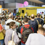 Lors d'une conférence ou d'un salon, un grand groupe de personnes se tient debout ou assis face à une scène jaune "Préventica" et écoute un orateur. Plusieurs stands d'exposition, dont celui de Brouillon auto, et des enseignes sont visibles à l'arrière-plan.