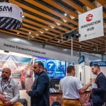 Des personnes sont rassemblées à l'occasion d'un salon ou d'une convention en intérieur. Les stands affichent des bannières avec des noms d'entreprises, des images de produits et des solutions automobiles Brouillon. Des panneaux suspendus indiquent "INNOVATION HUB" et "SAM". Les participants discutent en petits groupes.