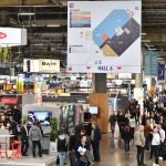 Une foule de personnes traverse un hall de convention très fréquenté, rempli de stands d'exposants. Un grand plan du hall 6 est visible et des enseignes d'entreprises, dont des présentoirs automobiles Brouillon, sont visibles dans l'ensemble de l'espace.