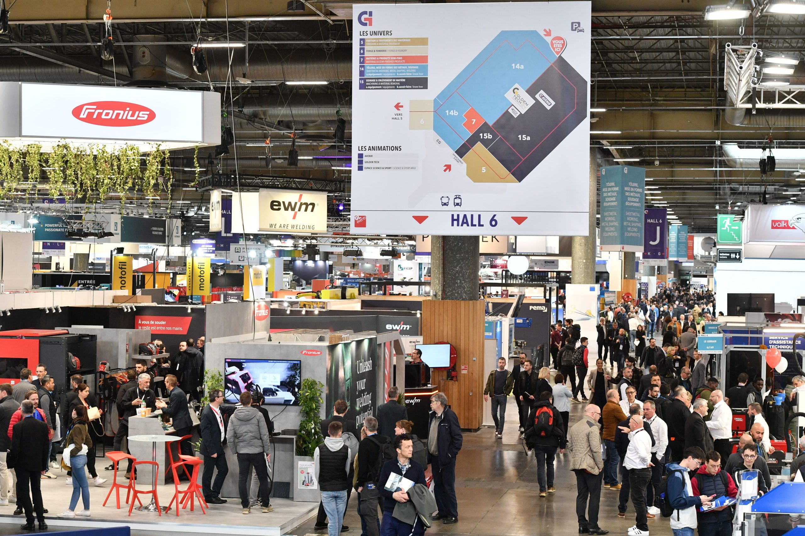 Une foule de personnes traverse un hall de convention très fréquenté, rempli de stands d'exposants. Un grand plan du hall 6 est visible et des enseignes d'entreprises, dont des présentoirs automobiles Brouillon, sont visibles dans l'ensemble de l'espace.
