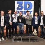 Neuf personnes se tiennent en rang à l'intérieur, certaines tenant des récompenses, devant un panneau portant l'inscription "3D PRINT". Vêtues d'une tenue décontractée et de badges de conférence, elles présentent les réalisations en matière d'impression 3D à l'aide d'objets imprimés en 3D exposés sur une table avant l'événement de 2026.