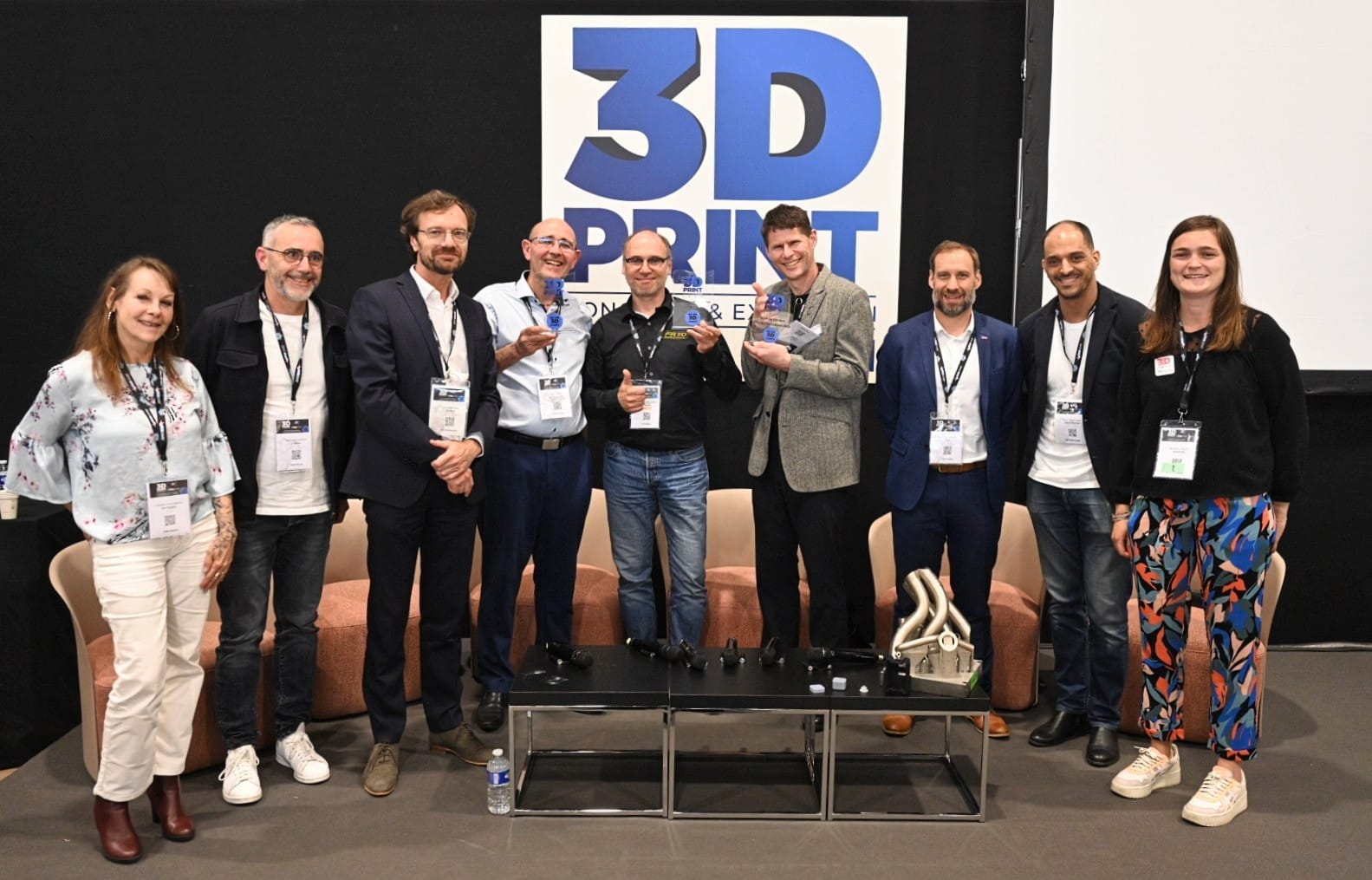 Neuf personnes se tiennent en rang, souriant à la caméra devant un panneau "3D PRINT". Certaines tiennent des objets, peut-être des objets imprimés en 3D ou des prototypes de Brouillon auto. Elles portent des vêtements d'affaires décontractés ou formels et des badges de conférence.