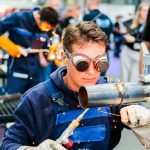 Une personne portant des lunettes de protection et des gants utilise une torche de soudage sur un tuyau en métal, faisant jaillir des étincelles - une scène de compétences et d'innovations pratiques. À l'arrière-plan, d'autres personnes travaillent dans un atelier animé, reflétant ainsi l'esprit de Schweissen & Schneiden édition 2025.