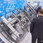 Plusieurs hommes en tenue d'affaires observent un grand composant de machine industrielle exposé verticalement dans un cadre métallique protecteur à l'EuroBLECH 2026, qui présente des innovations sur une toile de fond bleue et pointillée au salon professionnel.