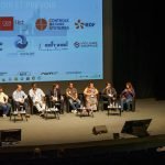 Un panel de neuf personnes est assis sur scène devant un grand écran projeté affichant divers logos d'entreprises, dont celui de Brouillon auto, sous le regard d'un public dans un auditorium sombre.