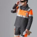 Une personne portant un uniforme de travail gris et orange à haute visibilité et des lunettes de soleil foncées tient un document enroulé sur un fond gris uni.