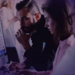 Trois personnes assises à un bureau regardent attentivement un grand écran d'ordinateur affichant des graphiques et des données. L'éclairage est faible et des silhouettes floues apparaissent à l'arrière-plan.