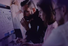 Trois personnes assises à un bureau regardent attentivement un grand écran d'ordinateur affichant des graphiques et des données. L'éclairage est faible et des silhouettes floues apparaissent à l'arrière-plan.