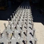 Des rangées de montants métalliques régulièrement espacés, fixés par soudage de goujons, sont attachés à une longue poutre métallique plate dans un atelier, la lumière du soleil projetant des ombres sur la surface métallique.