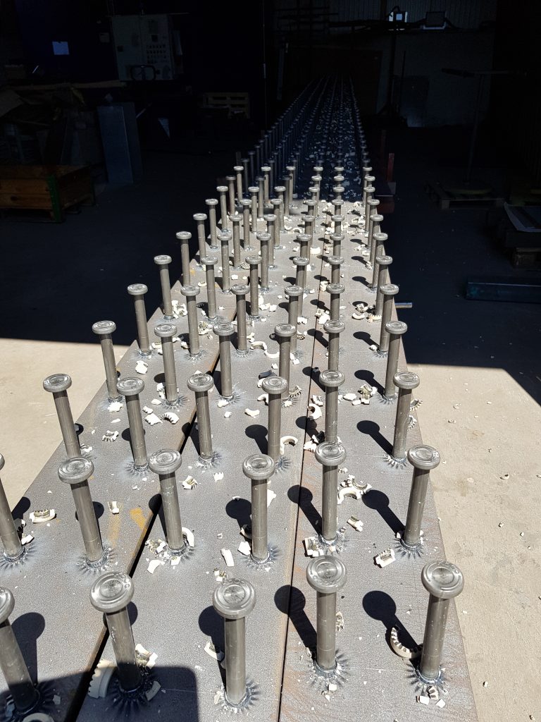 Des rangées de montants métalliques régulièrement espacés, fixés par soudage de goujons, sont attachés à une longue poutre métallique plate dans un atelier, la lumière du soleil projetant des ombres sur la surface métallique.