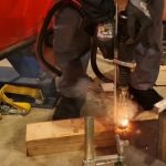 Une personne portant des gants et des vêtements de protection utilise un découpeur au plasma pour découper une feuille de métal. Des étincelles brillantes et de la fumée sont visibles. L'installation, idéale pour souder sans contraintes, comprend des pinces métalliques et des outils sur une plate-forme en bois.