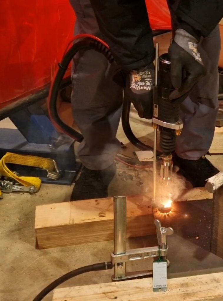 Une personne portant des gants et des vêtements de protection utilise un découpeur au plasma pour découper une feuille de métal. Des étincelles brillantes et de la fumée sont visibles. L'installation, idéale pour souder sans contraintes, comprend des pinces métalliques et des outils sur une plate-forme en bois.