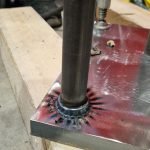 Un gros plan du soudage de goujons montre une tige de métal soudée à une plaque, avec des traces de brûlure noircies autour de la soudure. L'ensemble repose sur une planche en bois, avec des outils et le pied d'une personne visibles à l'arrière-plan.