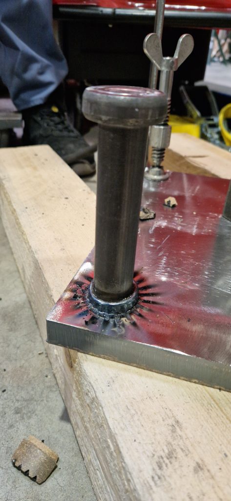 Un gros plan du soudage de goujons montre une tige de métal soudée à une plaque, avec des traces de brûlure noircies autour de la soudure. L'ensemble repose sur une planche en bois, avec des outils et le pied d'une personne visibles à l'arrière-plan.