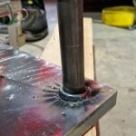 Une tige métallique est soudée verticalement sur une plaque métallique plate qui repose sur du bois. La soudure et la zone environnante présentent des marques de soudage de goujons. Le pied d'une personne et des outils sont visibles à l'arrière-plan.