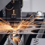 Vue rapprochée d'une machine industrielle de découpe au laser en fonctionnement, émettant des étincelles lumineuses lors de la découpe d'une feuille de métal sur une ligne de production.