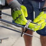 Une personne portant des gants de travail Helix 3014IMP jaune vif assemble des barres d'armature en acier avec des attaches en fil de fer, préparant probablement le renforcement de la construction en béton. Seuls les mains et le bas du corps sont visibles, mettant en avant la sécurité et la protection à l'extérieur.
