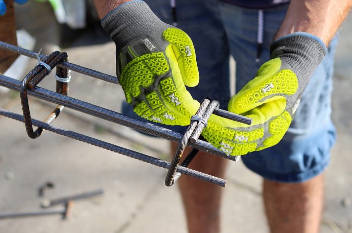 Une personne portant des gants de travail Helix 3014IMP jaune vif assemble des barres d'armature en acier avec des attaches en fil de fer, préparant probablement le renforcement de la construction en béton. Seuls les mains et le bas du corps sont visibles, mettant en avant la sécurité et la protection à l'extérieur.