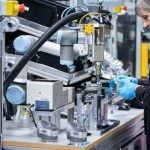 Une personne portant des gants bleus manipule des machines ou des bras robotisés sur un établi dans un environnement de fabrication ou de laboratoire. Divers composants et équipements mécaniques sont visibles sur la table.