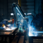 Une personne effectue un soudage sur un établi tandis qu'un bras robotisé Universal Robots UR8 Long fonctionne à proximité, tous deux entourés d'étincelles et de fumée, dans un environnement industriel faiblement éclairé.