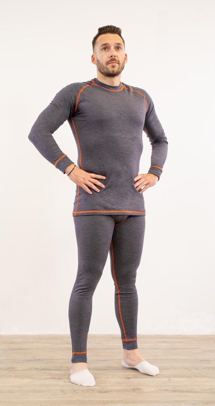 Un homme se tient debout sur un sol en bois, vêtu de sous-vêtements gris assortis aux coutures orange et de chaussettes blanches. Ses mains sont posées sur ses hanches, le regard légèrement tourné vers le haut, évoquant la confiance et la sécurité sur le fond blanc uni.