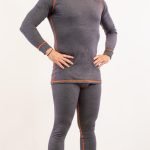 Un homme se tient à l'intérieur, vêtu de sous-vêtements techniques gris à manches longues assortis, avec des coutures orange. Les mains sur les hanches et le regard légèrement tourné vers le haut, il associe l'ensemble à des chaussettes blanches, prêt pour le confort et la sécurité dans n'importe quelle activité.