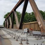 Un pont en acier en construction présente des poutres métalliques apparentes et de nombreux soudages de goujons, avec des tiges d'acier dépassant du béton. Un générateur et du matériel sont posés sur le pont, tandis que les arbres et le ciel forment l'arrière-plan.