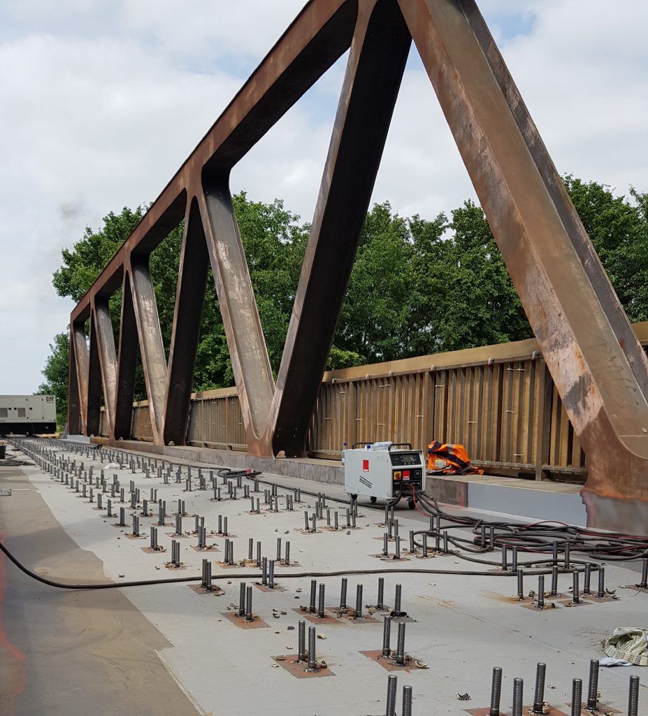 Un pont en acier en construction présente des poutres métalliques apparentes et de nombreux soudages de goujons, avec des tiges d'acier dépassant du béton. Un générateur et du matériel sont posés sur le pont, tandis que les arbres et le ciel forment l'arrière-plan.