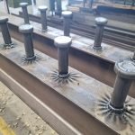 Vue rapprochée de goujons de cisailles métalliques soudés sur une poutre d'acier dans un atelier, mettant en évidence la précision du soudage de goujons. Les goujons présentent des marques de soudure visibles à leur base, tandis que d'autres poutres métalliques sont empilées à l'arrière-plan.