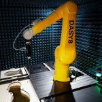 Un bras robotique jaune étiqueté "DASY8" fait une démonstration de robotisation dans une chambre anéchoïque, entourée de panneaux de mousse absorbant le son et d'équipements. Un petit voyant lumineux est visible sur le côté droit de la table.